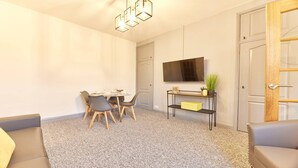 A modern living room with a dining area, a flat-screen TV mounted on the wall, a small plant on a shelf, and a door with a wooden frame. - Host Stay Inness House (Bedlington)
