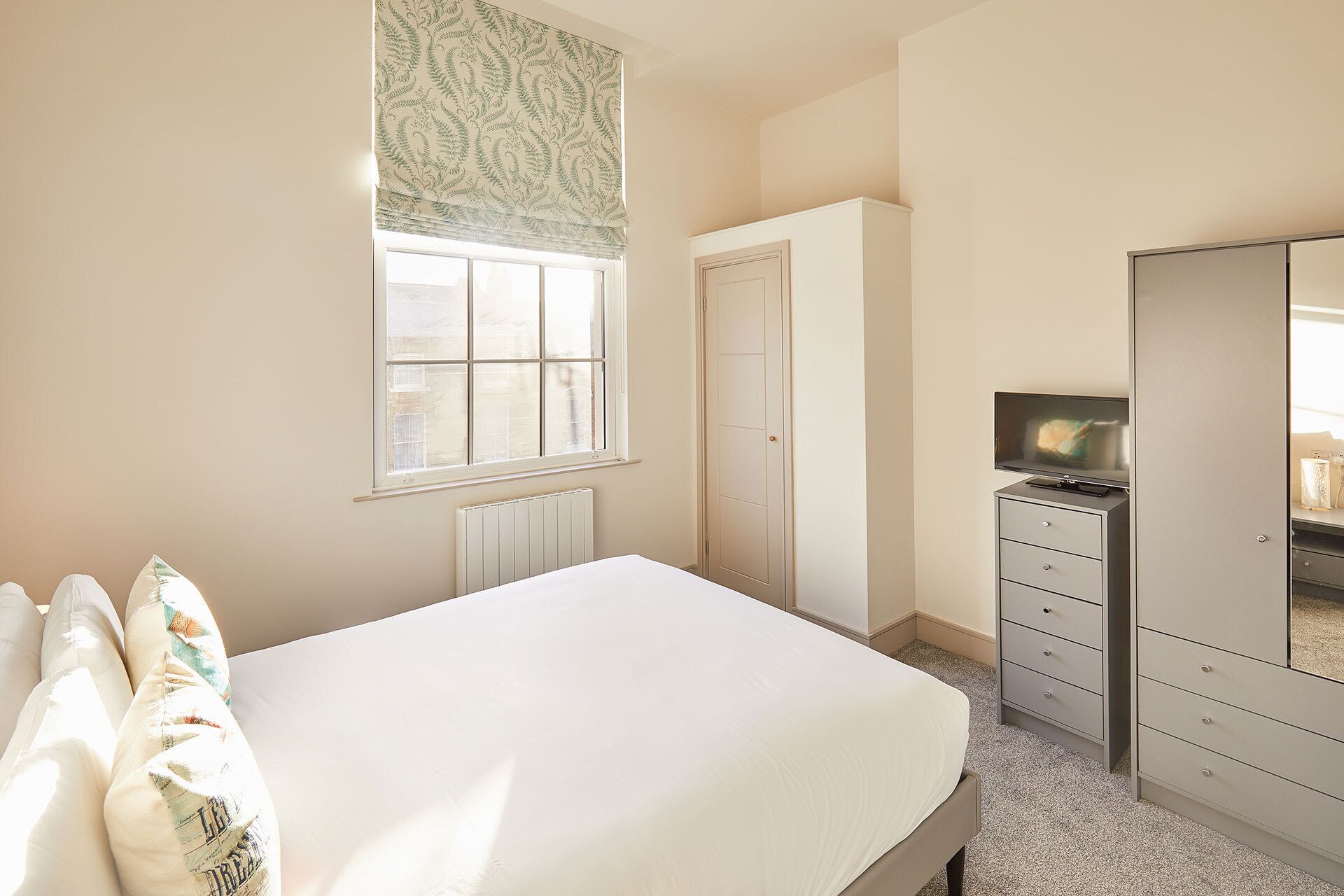 Un dormitorio con cama, cómoda, espejo, ventana con cortina estampada y radiador.