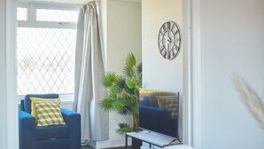 Un salon avec un canapé bleu, un téléviseur posé sur un meuble, une grande plante et une horloge décorative accrochée au mur.