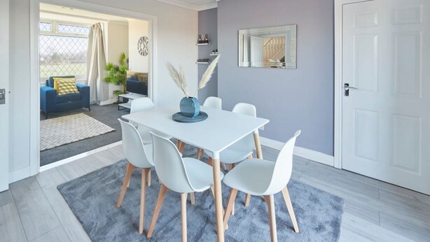 Un coin repas avec une table et des chaises blanches, un tapis gris et un vase bleu contenant des plumes d’herbe des pampas.