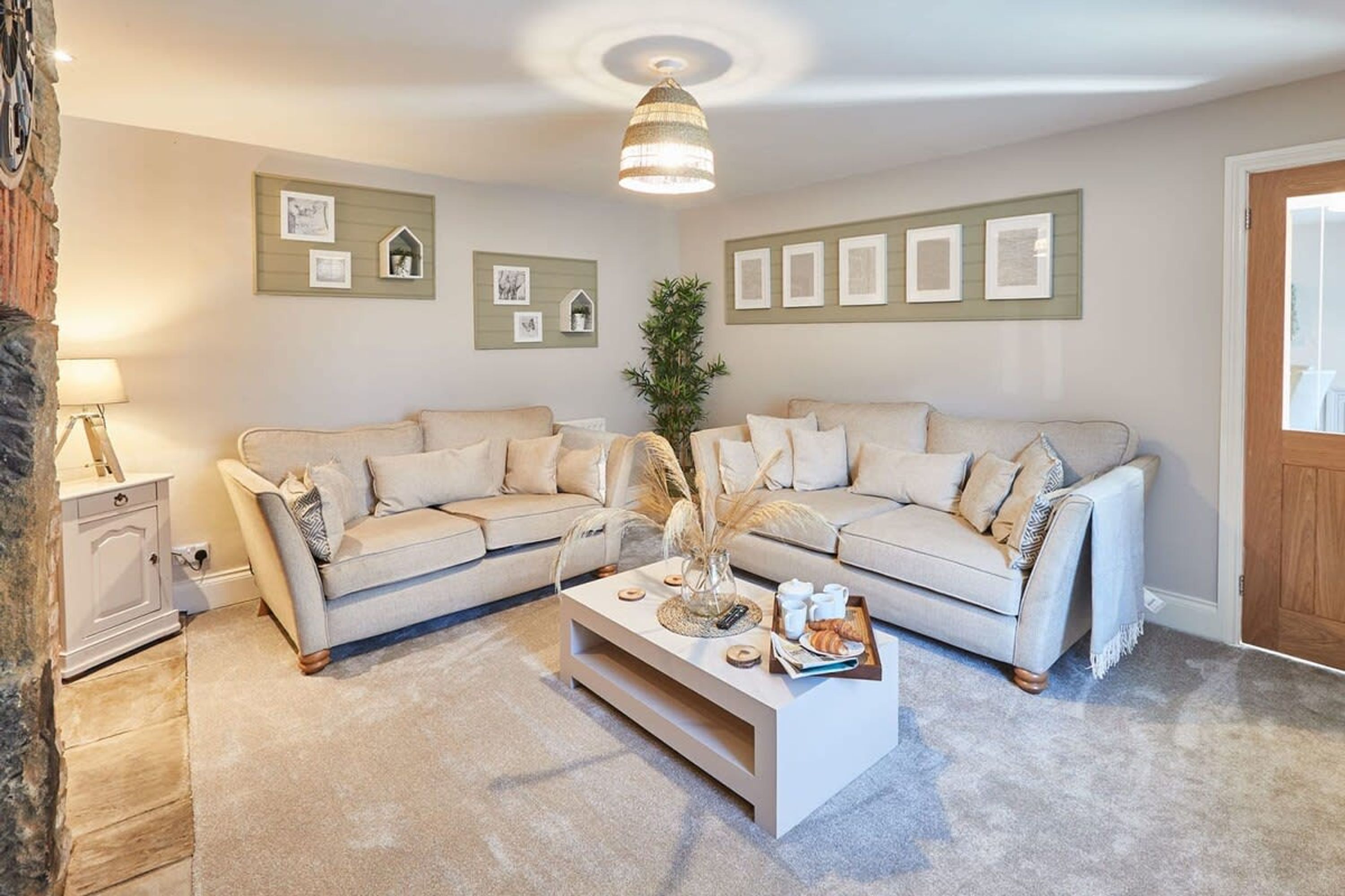 Ein Wohnzimmer mit einem beigefarbenen Ecksofa, einem weißen Couchtisch und einer Lampe. An der Wand hängen gerahmte Bilder und in der Ecke steht eine Pflanze.