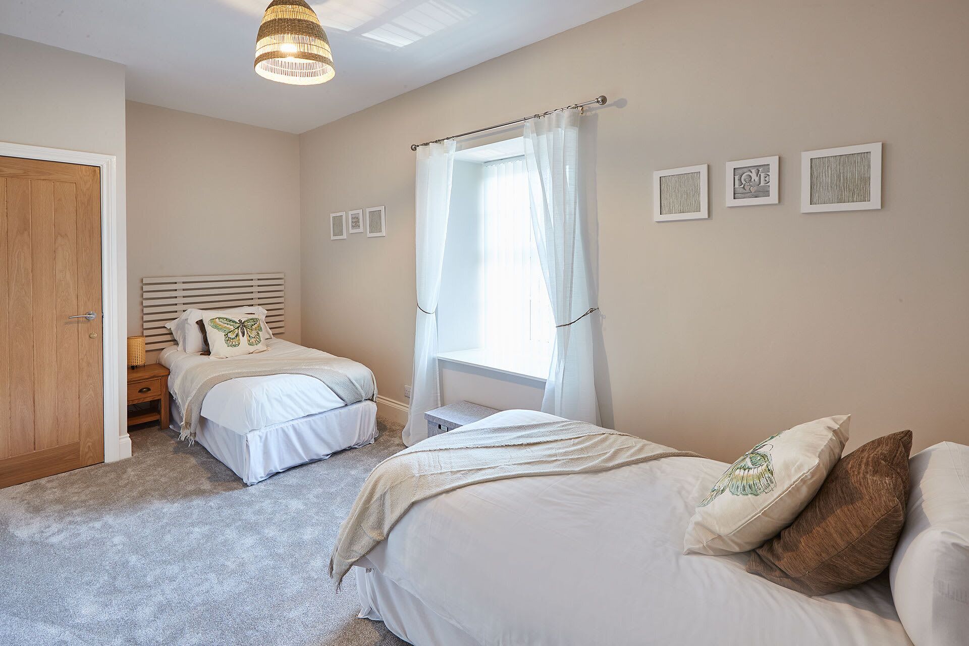 A bedroom with two beds, a wooden door, a window with sheer curtains, and wall art.