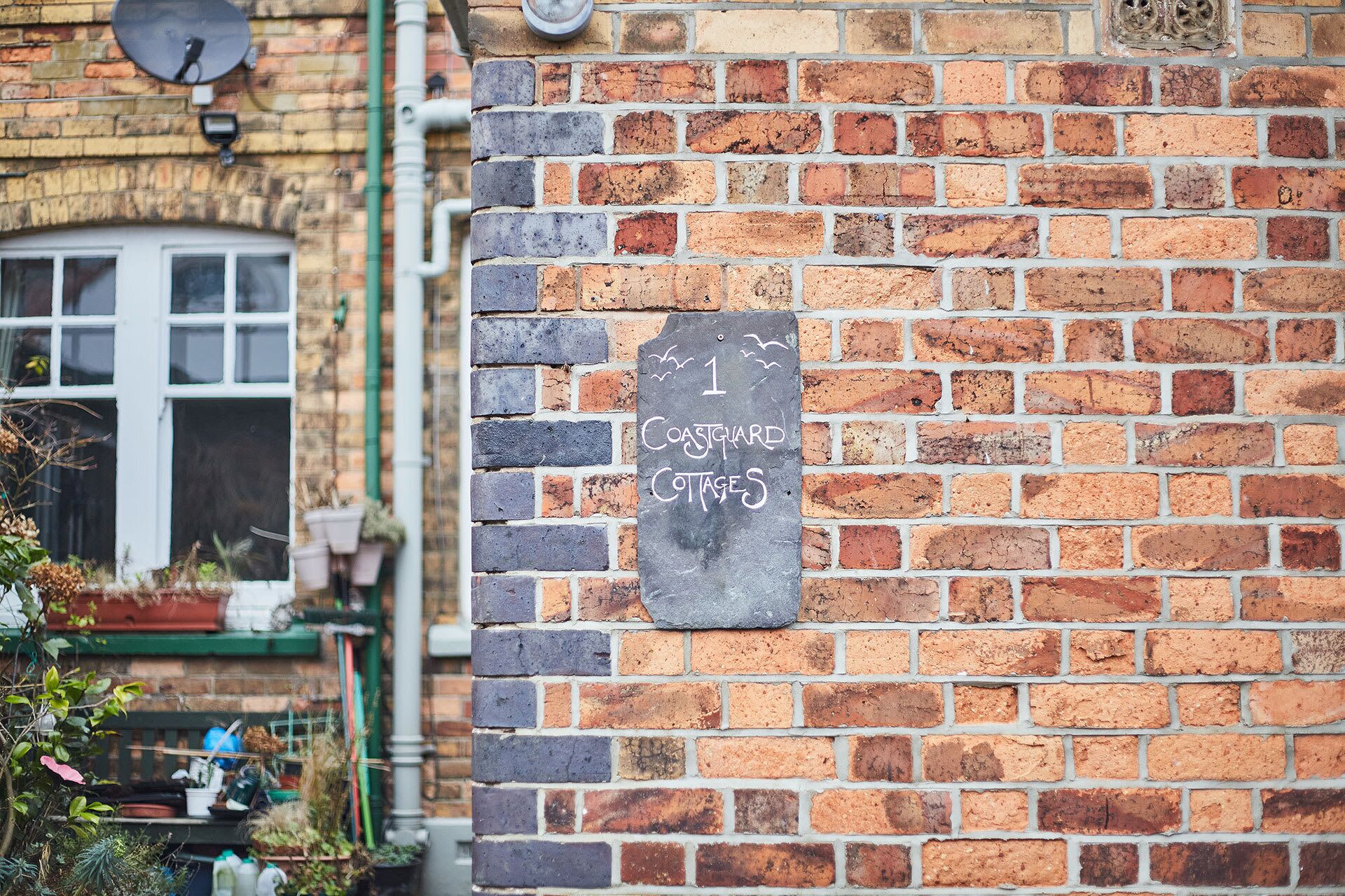 A brick wall with a chalkboard sign indicating '1 Coastguard Cottages'.