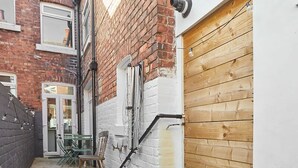 A narrow alleyway with a wooden bench, a chair, and a table set up against a brick wall. There is a wooden door on the right and a white wall with a window on the left. - Host Stay Upleatham Street (Saltburn-by-the-Sea)