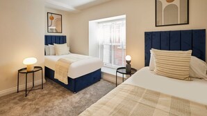 A hotel room with two beds, a large window with blinds, a framed artwork above the beds, and a small table with a lamp. - Host Stay 3 The Mount (Whitby)
