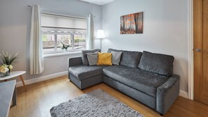 A modern living room with a grey sectional sofa, a small side table with a vase, a grey rug, a window with curtains, and a painting on the wall. - Host Stay Britton Hall Bungalow (Bishop Auckland)