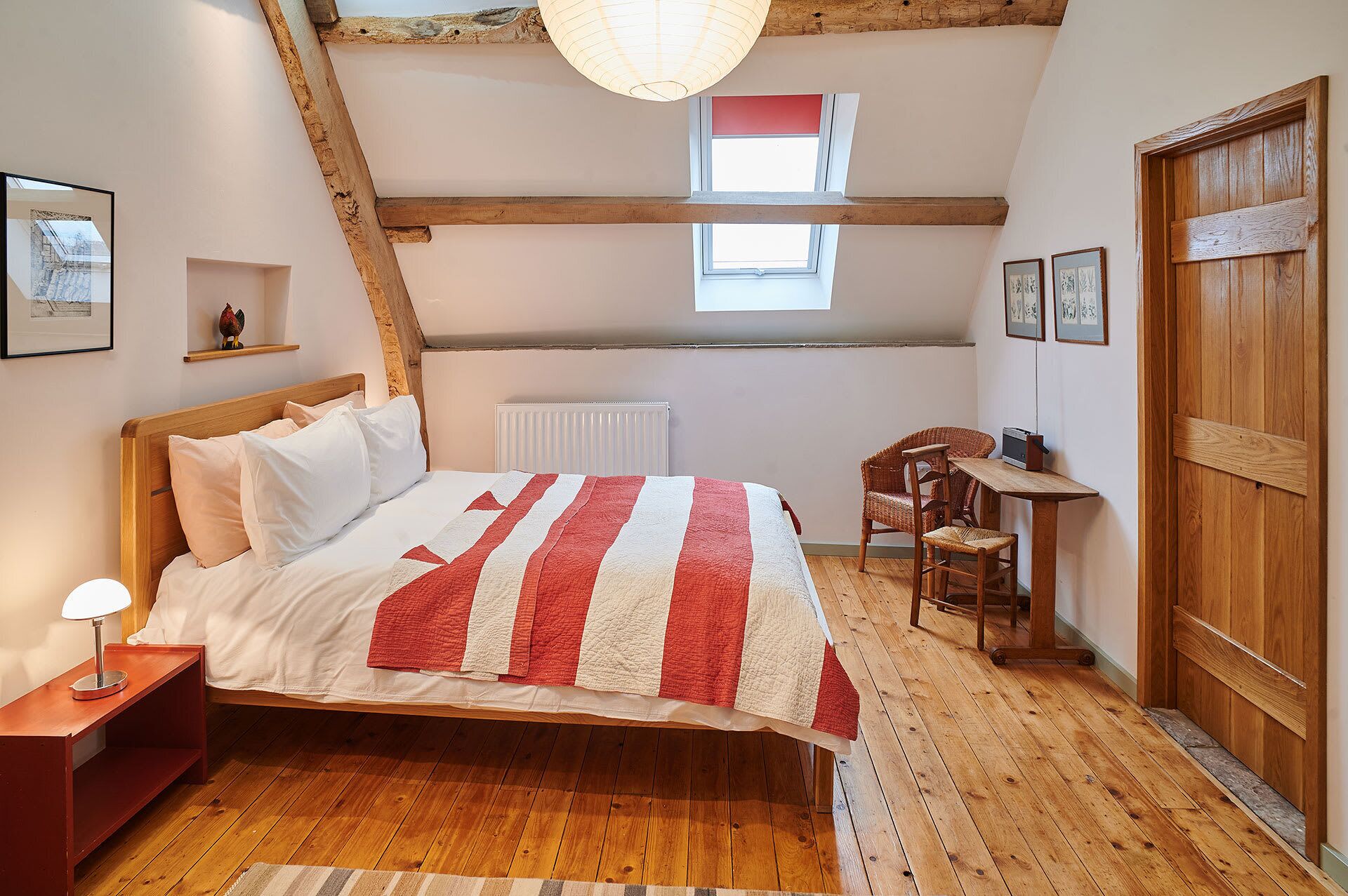 Et soveværelse med en træseng, et lille bord med en stol, en radiator og et vindue med rød gardin.
