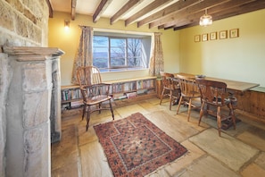 Une pièce chaleureuse avec un mur en pierre, des étagères en bois, une grande fenêtre avec des rideaux, un tapis à motifs et une table en bois avec des chaises.