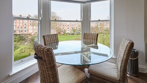 Ein Erker mit einem Glastisch und Korbstühlen, mit Blick auf einen Garten und Gebäude.