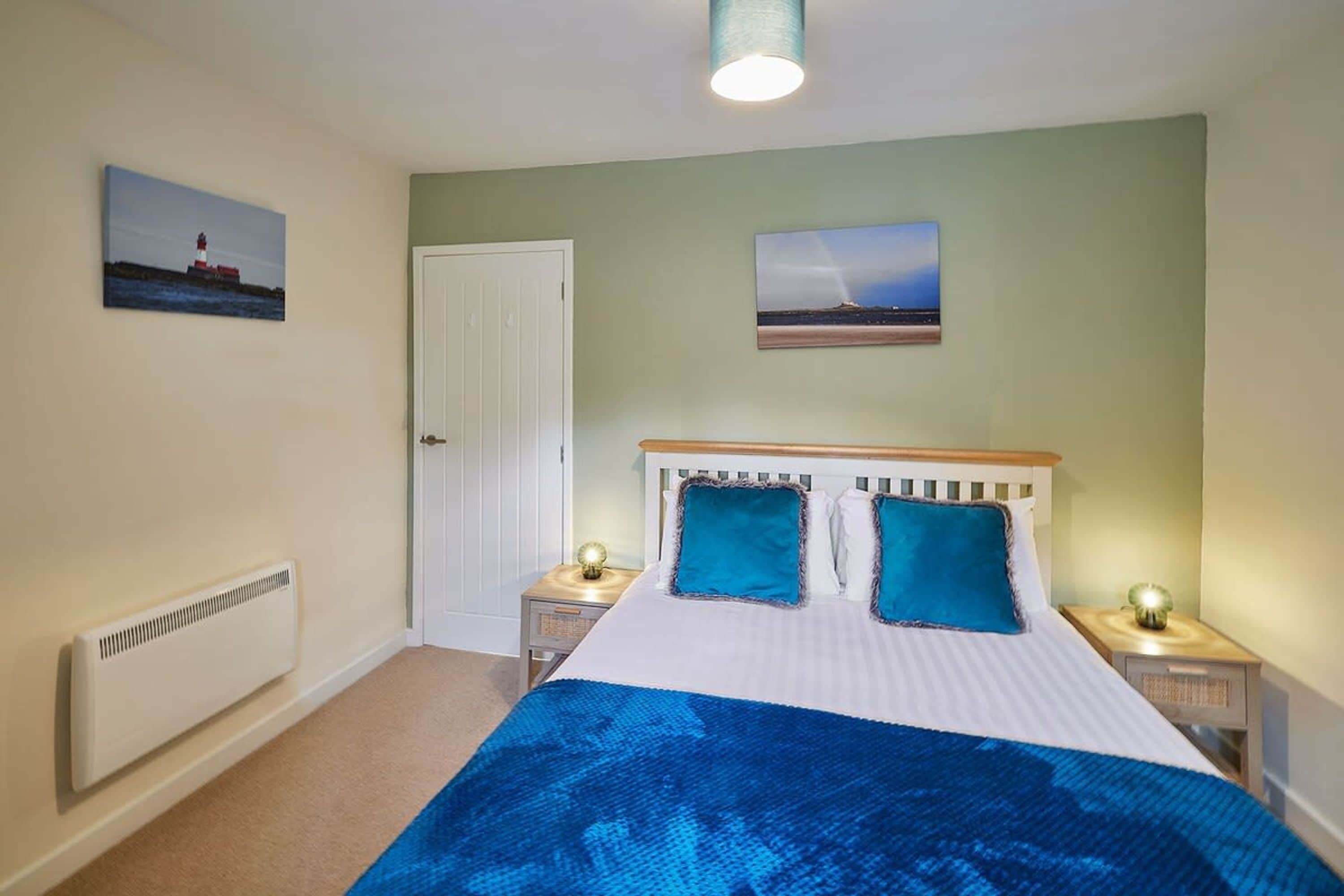 A bedroom with a bed, two bedside tables, a radiator, a door, and two wall-mounted pictures.