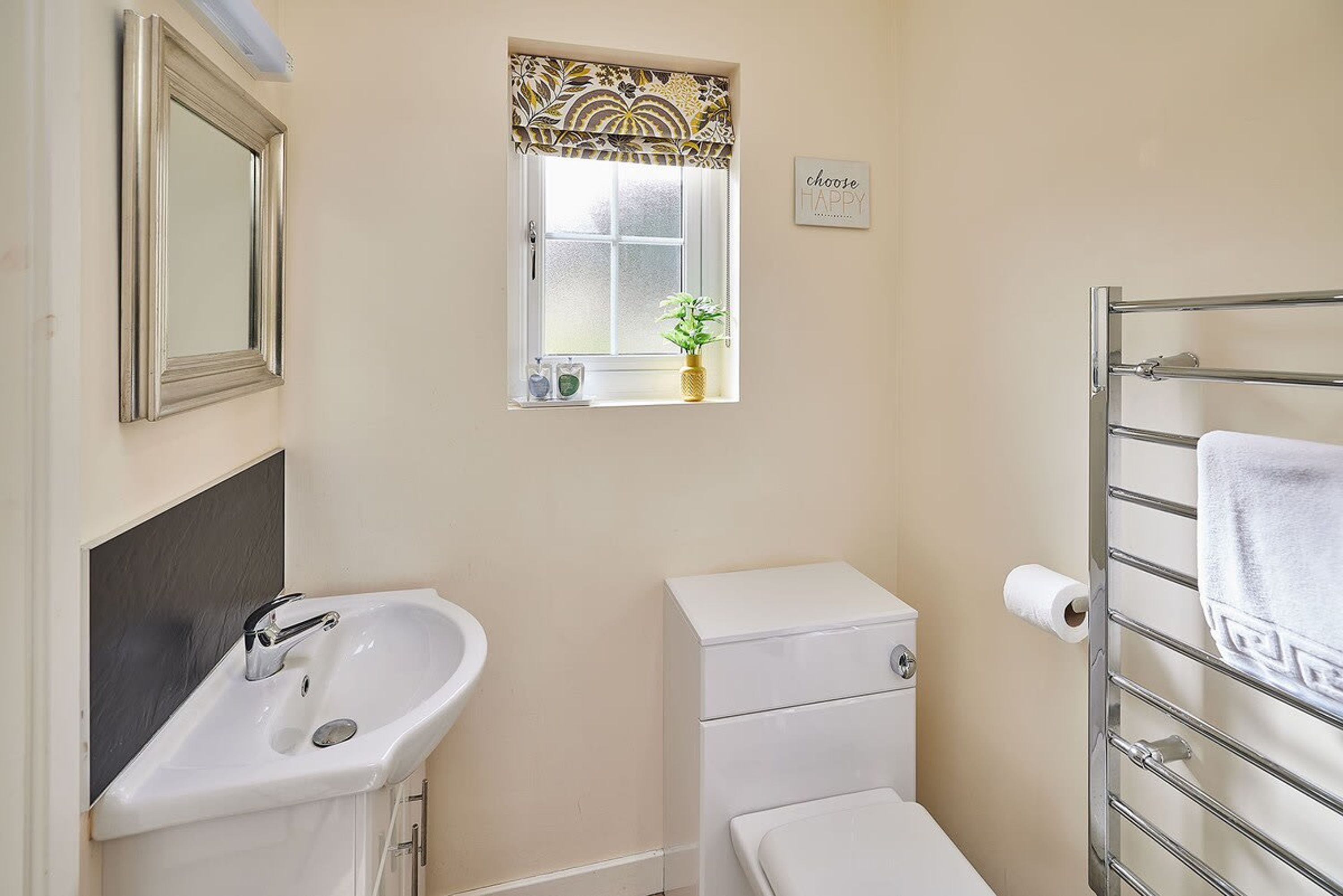 A small bathroom with a white sink, a toilet, a window with a decorative valance, a towel rack, and a sign that says "choose happy".