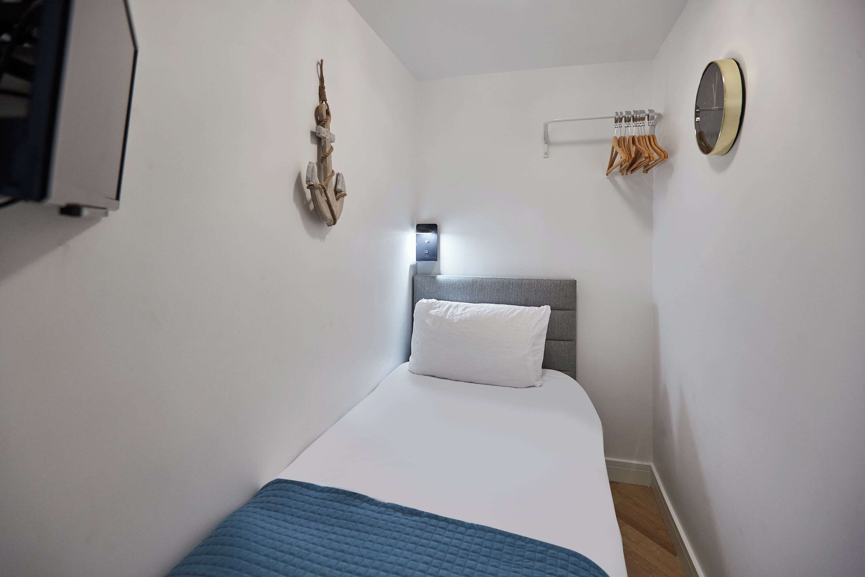 Ein kleines, ordentlich eingerichtetes Schlafzimmer mit einem Einzelbett, einem Nachttisch, einem Wandheizkörper, einem Kleiderständer und einem runden Spiegel.