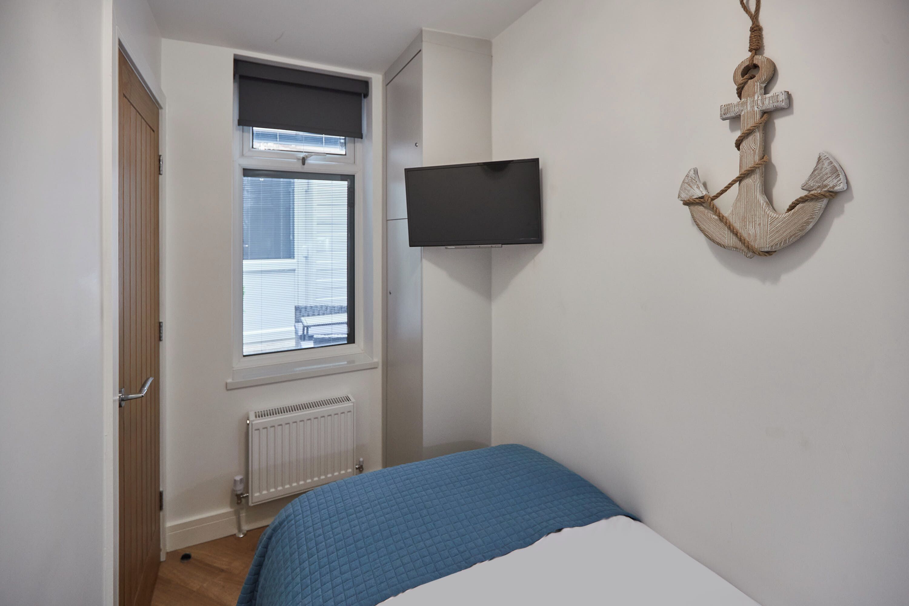 A bedroom with a bed, a window with blinds, a mounted TV, a radiator, and a wall-mounted anchor decoration.