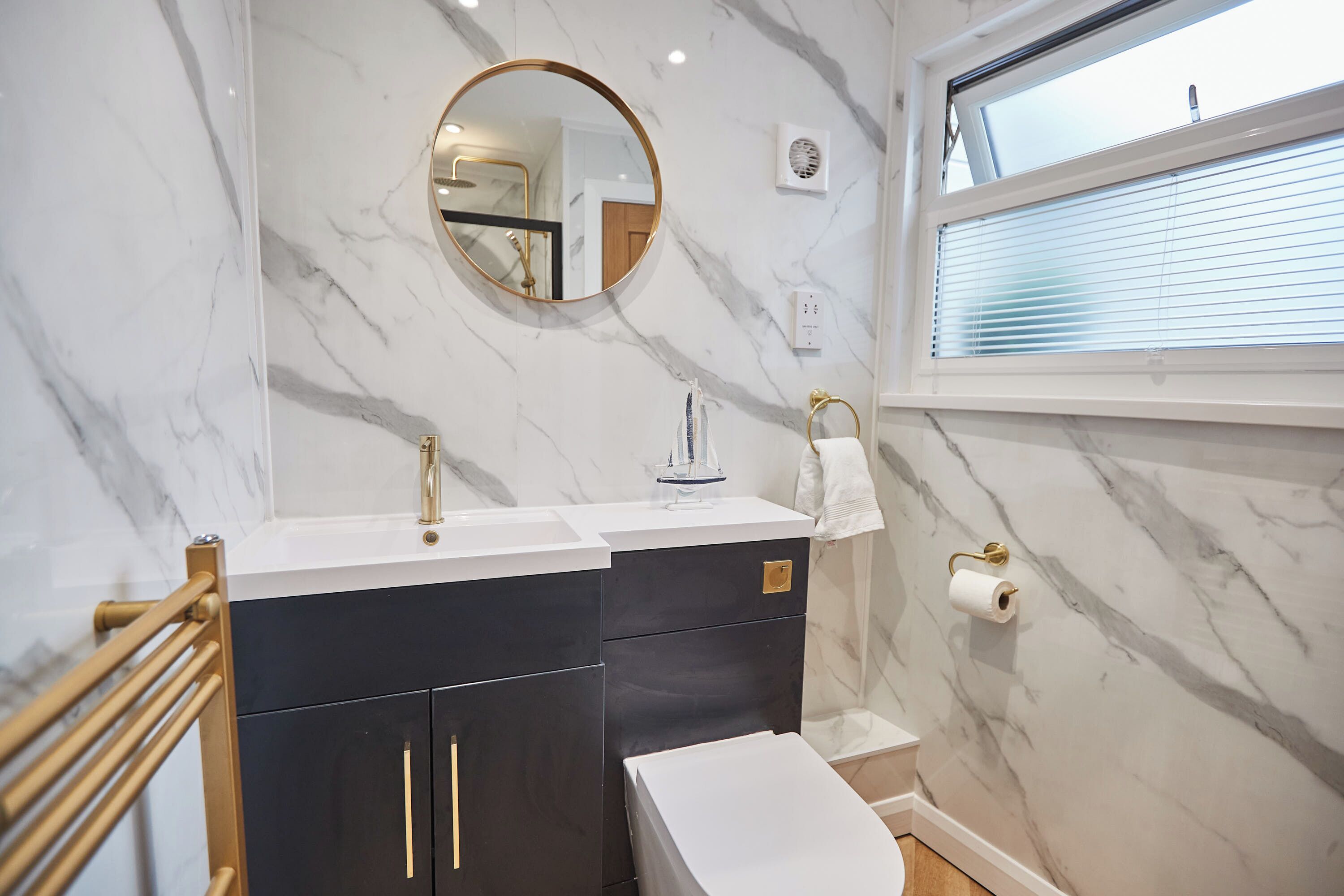 Une salle de bain avec un mur en marbre, un miroir rond, un lavabo, des toilettes et une fenêtre avec des stores.