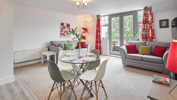 A modern living room with a dining area, grey sofas, a glass table, and red floral curtains. - Host Stay The Paddock (Bedlington)