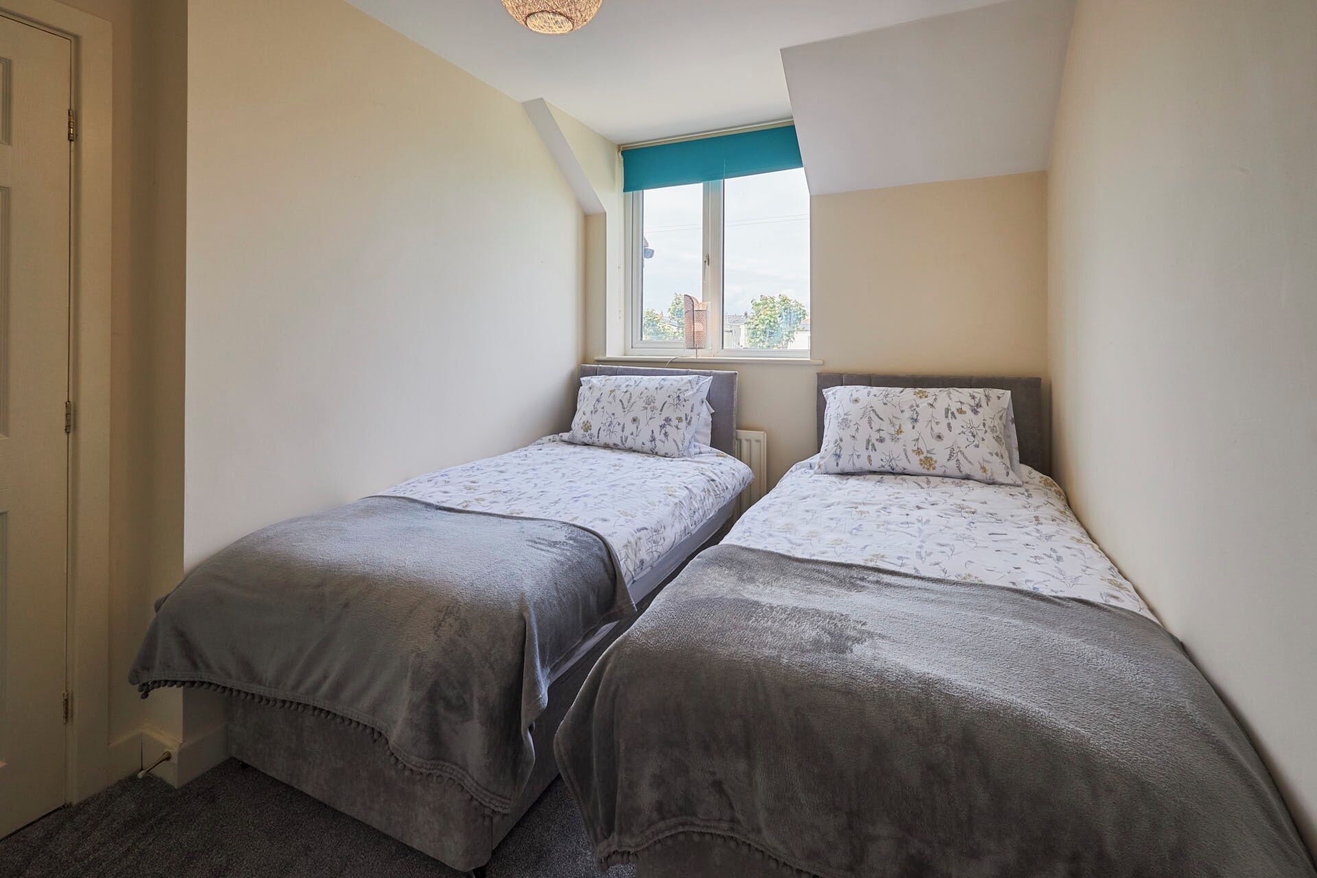 Une petite chambre à coucher, de taille modeste, avec deux lits simples, une fenêtre avec un store bleu et une porte située sur le côté gauche.