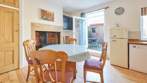 A dining area with a round table and wooden chairs, a brick fireplace, and a kitchen with a refrigerator and dishwasher. - Host Stay The Cottage in Corbridge (Corbridge)