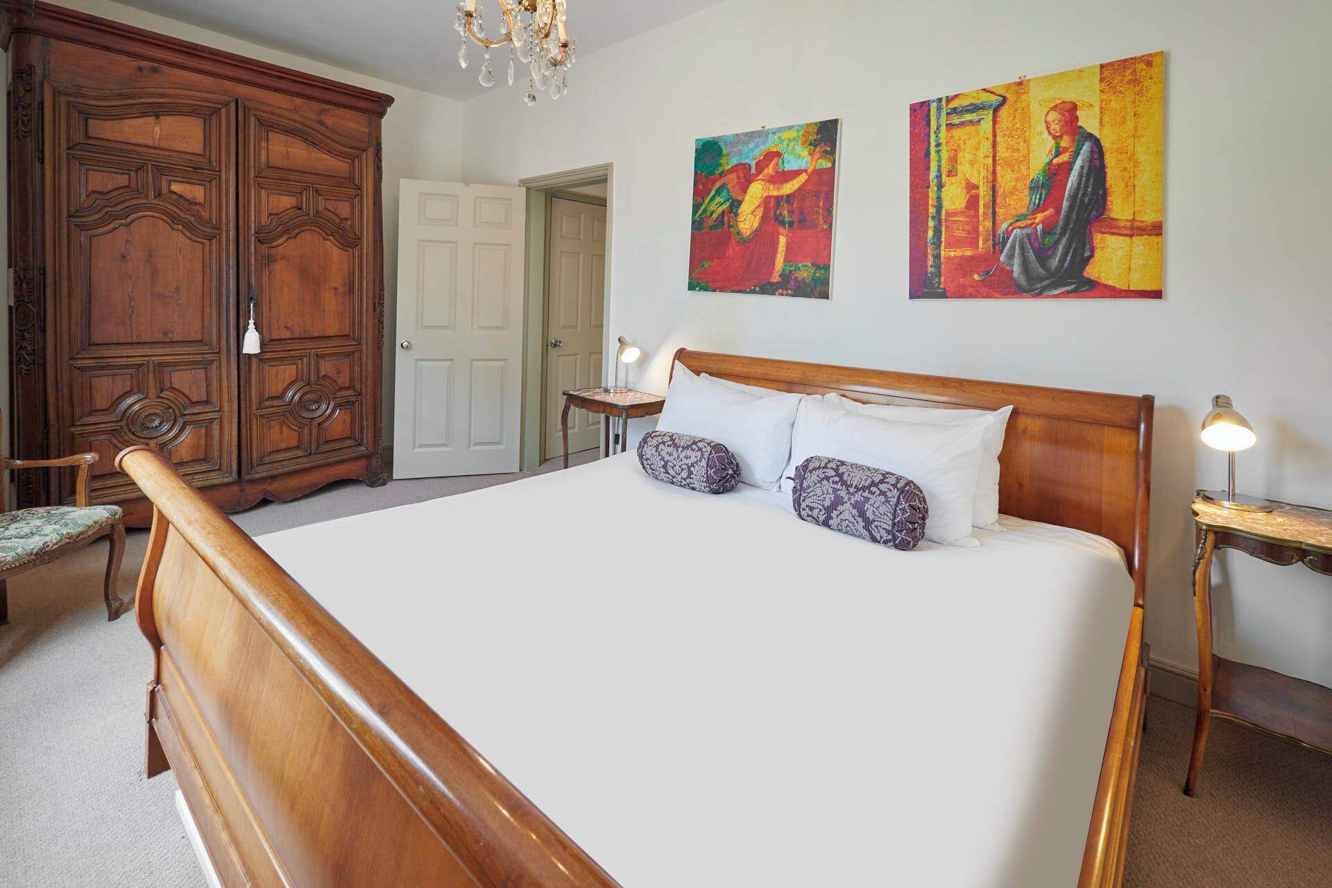 A bedroom with a large wooden bed, two paintings on the wall, a wardrobe, and a chandelier.