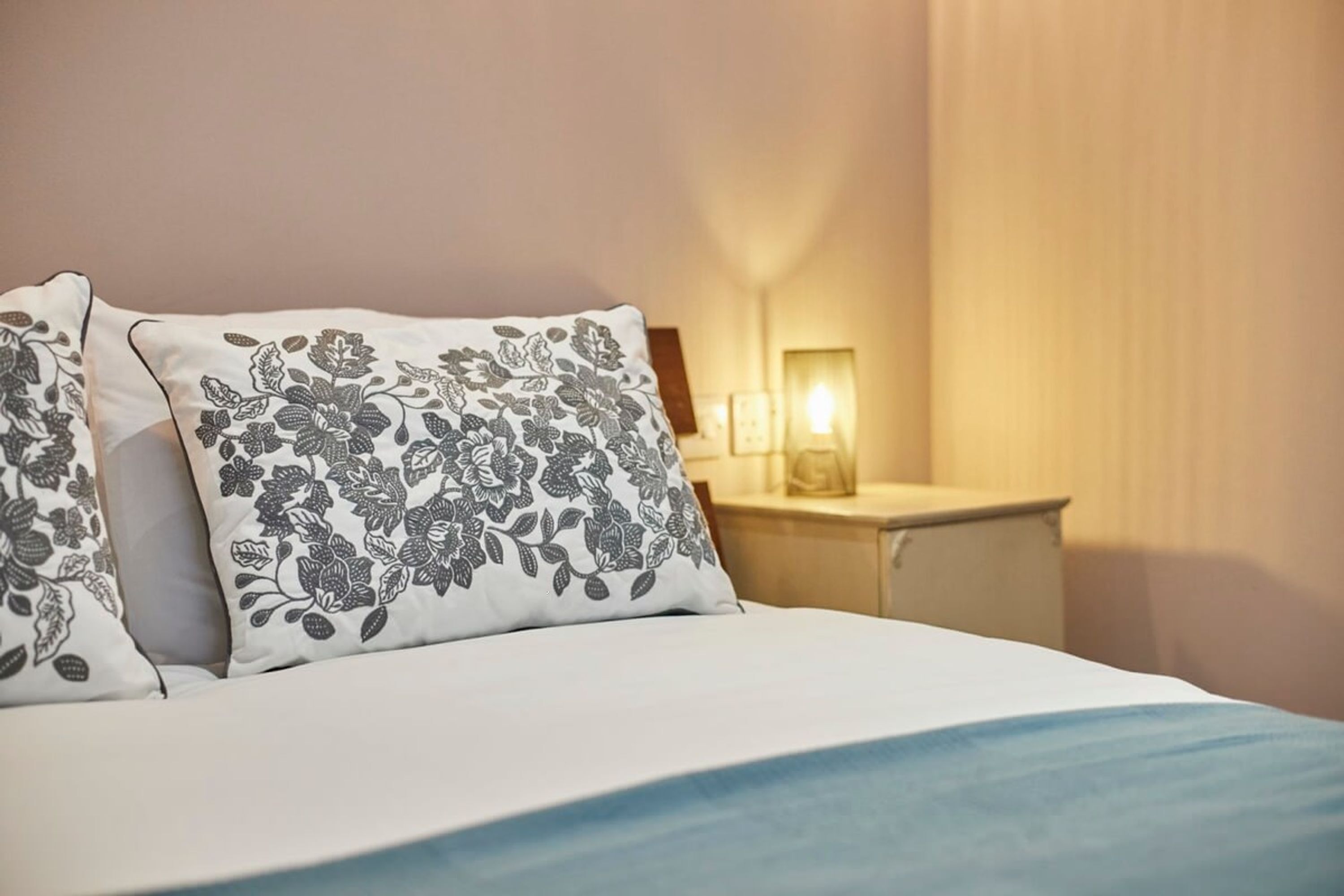 A neatly made bed with floral-patterned pillows, a bedside table with a lamp, and a wall-mounted light.