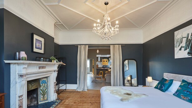 A bedroom with a large bed, a fireplace, a chandelier, and a view into the kitchen. - Host Stay Northville Arrifana (Whitley Bay)