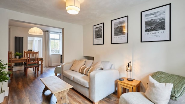 A modern living room with a sofa, a coffee table, and a dining area with a table and chairs. There are framed pictures on the wall and a lamp on a side table. - Host Stay Church View Cottage (Matlock)