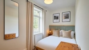 A bedroom with a bed, a wooden bedside table, a mirror, a window with curtains, and two framed pictures on the wall. - Host Stay Church View Cottage (Matlock)