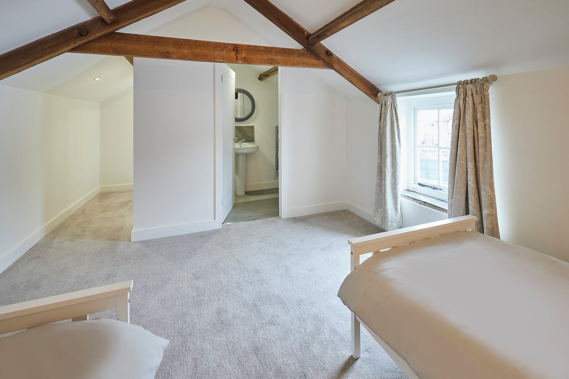 A bedroom with a sloped ceiling, a window with curtains, a small bathroom with a sink, and a bed.