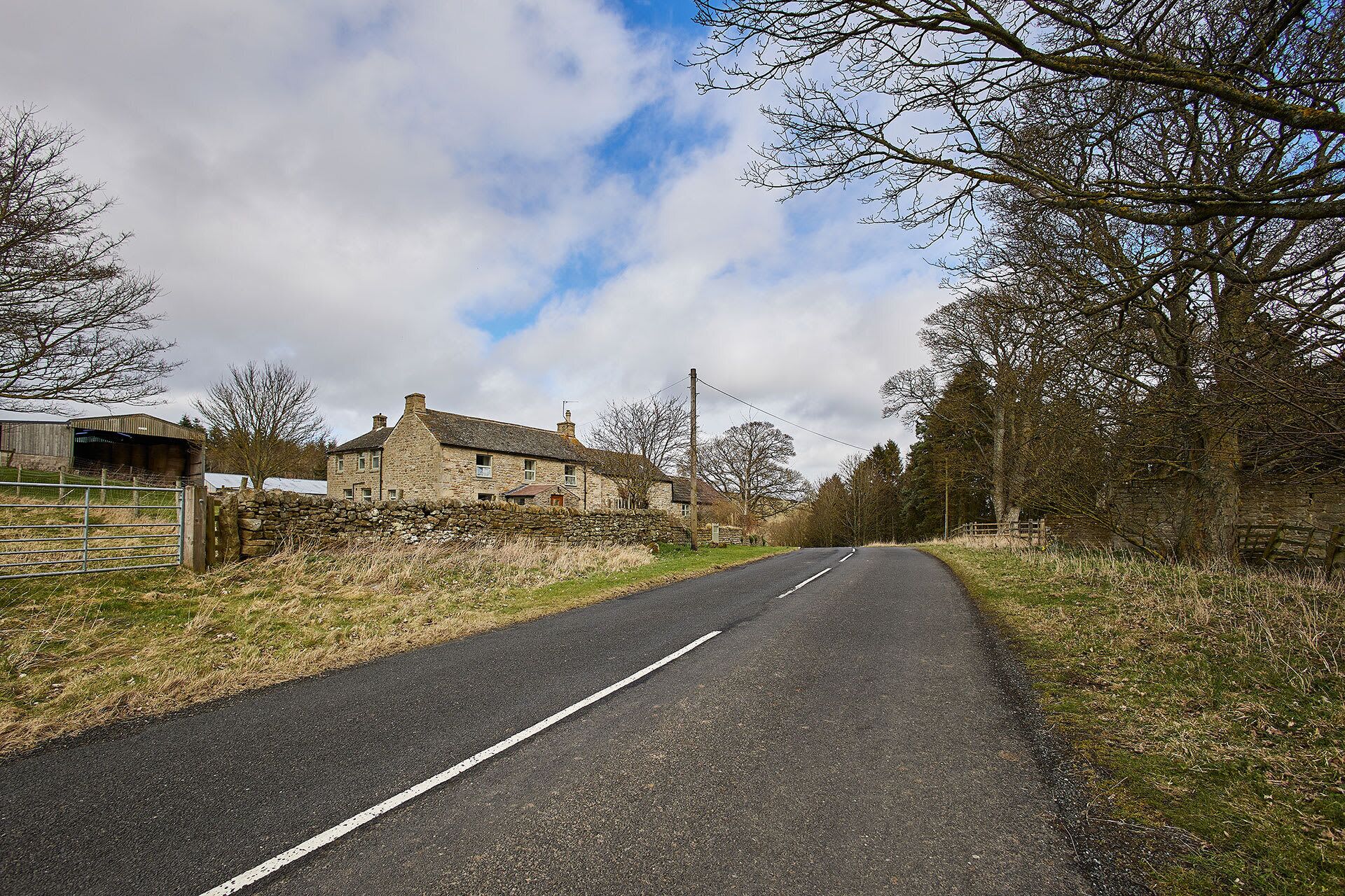 Eine ländliche Straße, begrenzt von Steinmauern und kahl stehenden Bäumen, führt zu einem Haus mit steinernen Wänden und einem geneigten Dach.