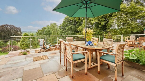 Ein runder Holztisch mit Stühlen, ein grüner Sonnenschirm und ein Teekessel auf einer Terrasse mit Steinboden und Blick auf Grünflächen.