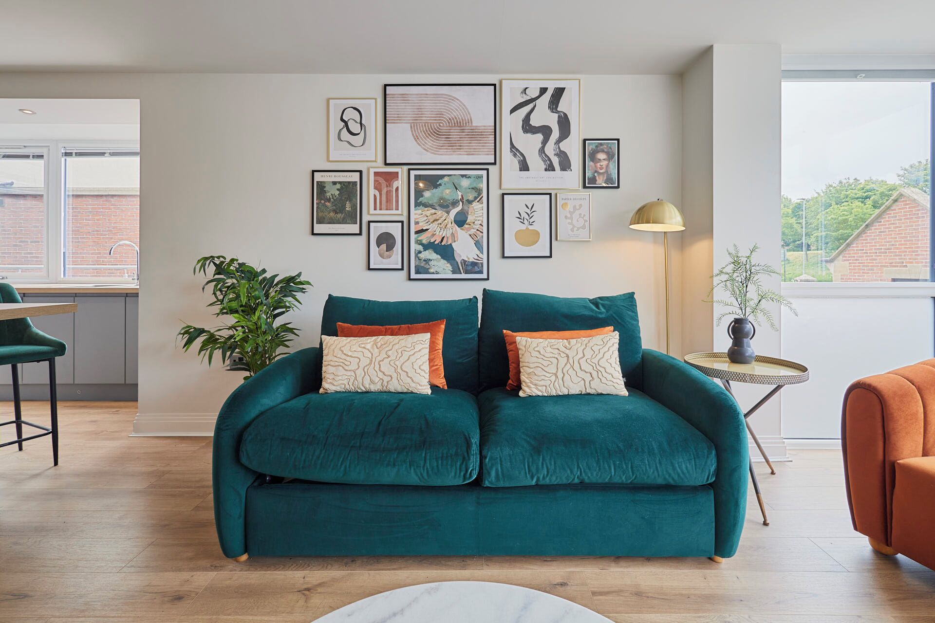 A teal sofa with patterned cushions, a small side table with a lamp, and a wall with various framed artworks.
