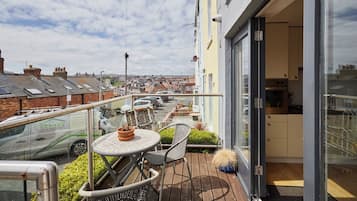 Een balkon met een tafel, stoelen, een bloempot en een uitzicht op de straat en gebouwen op de achtergrond.