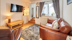 A living room with a brown leather sofa, a wooden coffee table, a TV mounted on the wall, a small table with a lamp, and a window with curtains. - Host Stay The Hideaway (Scarborough)