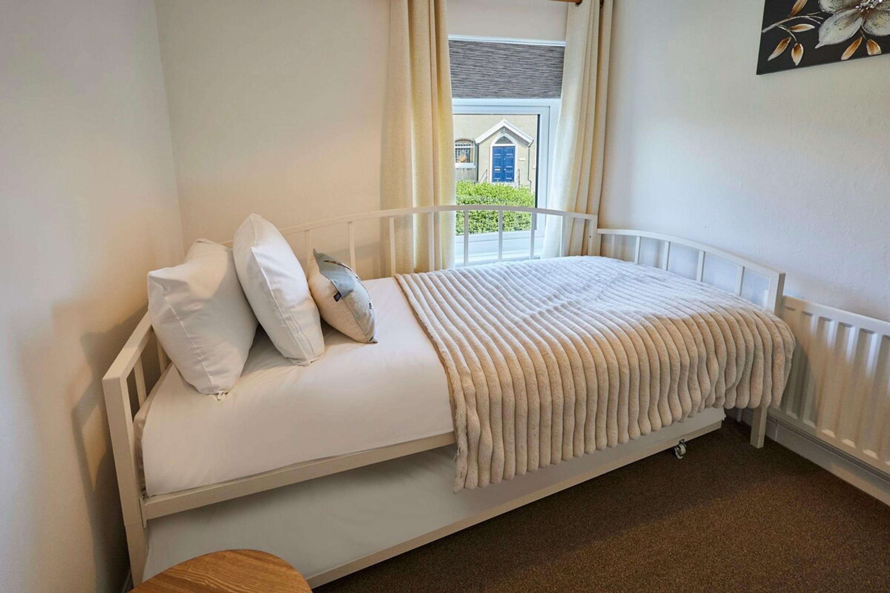 A single bed with white bedding and a beige throw blanket, a window with curtains, and a view of a house outside.