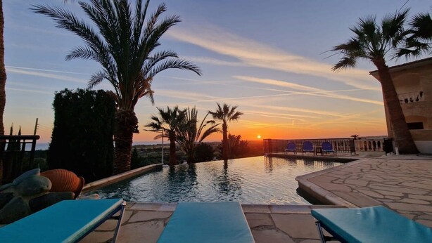 Zona de piscina con sillones de descanso, palmeras y vistas al atardecer.