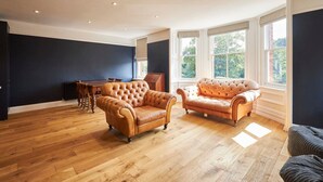 A spacious living room with a wooden floor, two leather sofas, a dining table, and large windows with a view of greenery. - Host Stay Fauna (Whitby)