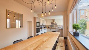 A modern kitchen with a wooden table, white cabinets, and a large window with a view of buildings. - Host Stay Darcey House (Scarborough)