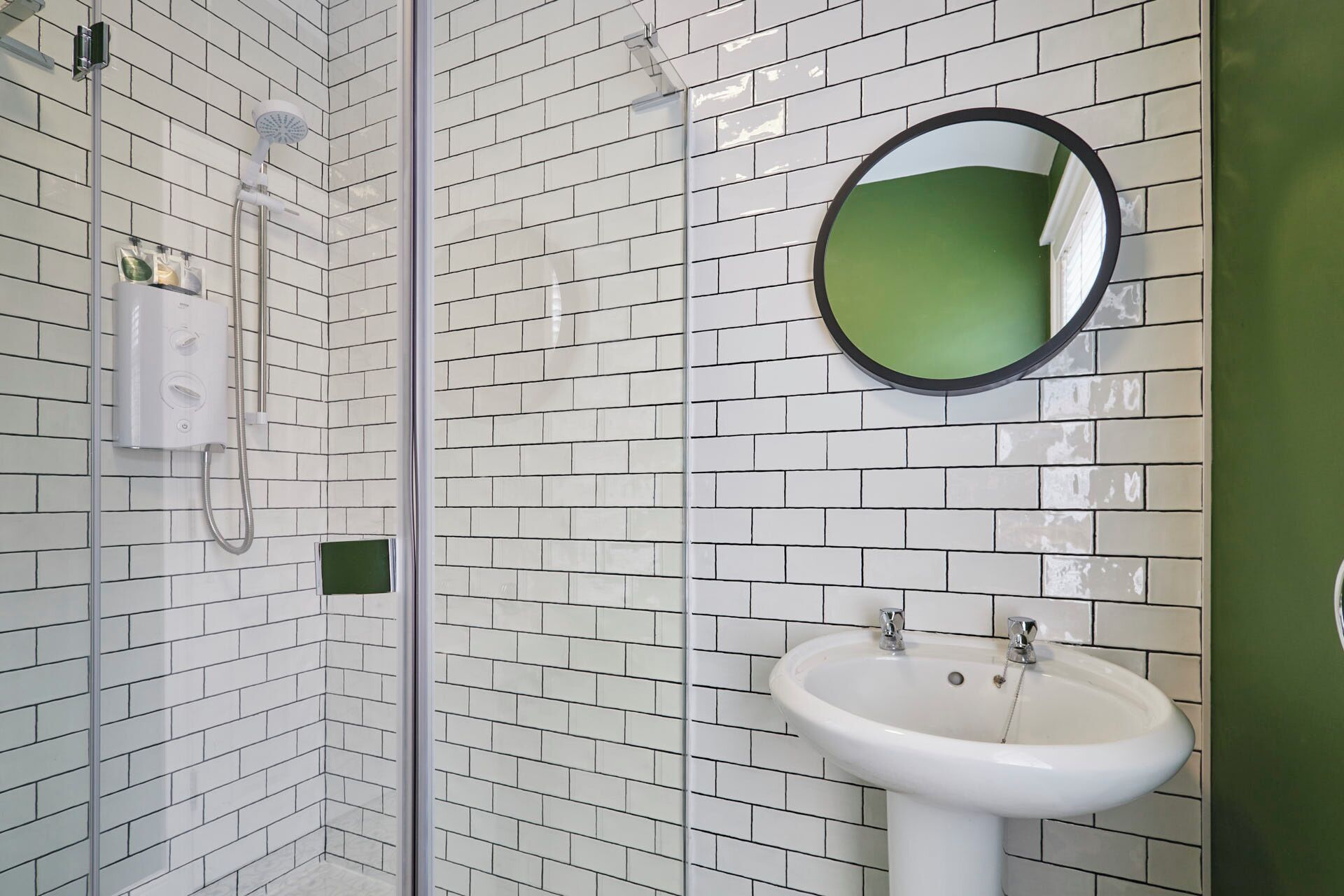 A bathroom with white subway tiles, a white sink, a round mirror, and a handheld showerhead.