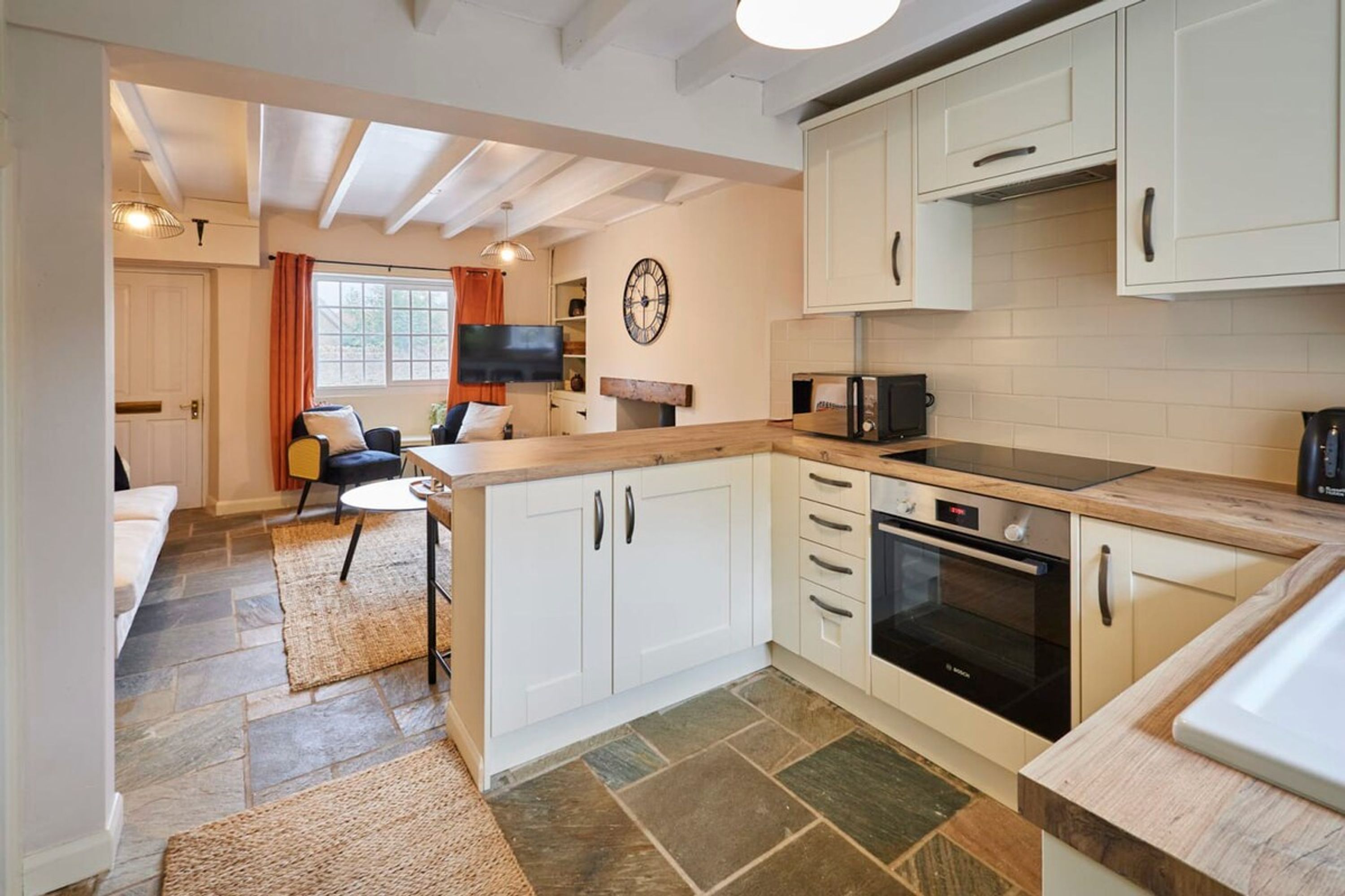 A modern kitchen with white cabinets, a built-in oven, and a microwave. There is a dining area with a table and chairs, and a living room with a sofa and a TV.