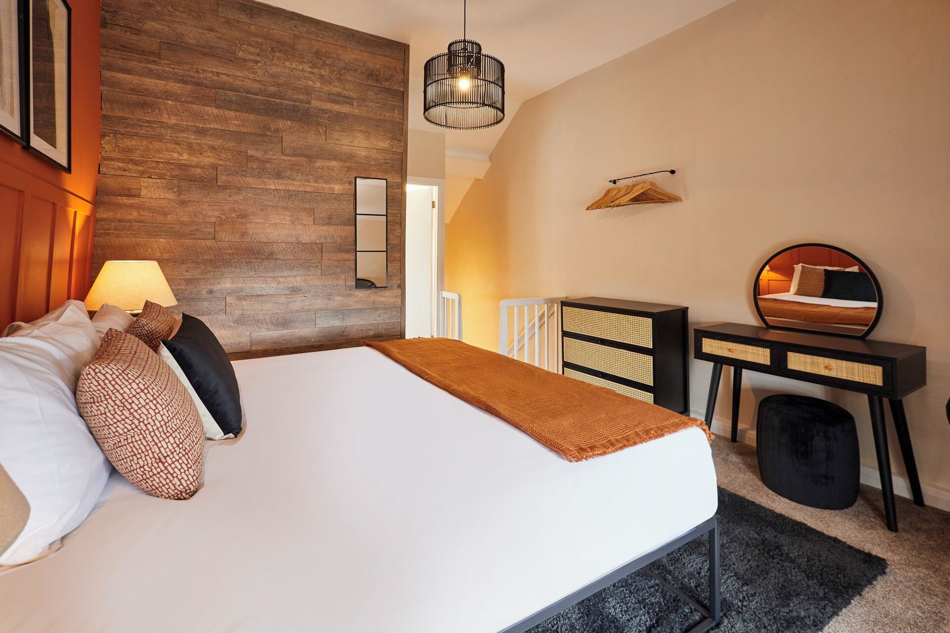 A neatly arranged hotel room with a bed, bedside lamp, a desk with a mirror, and a hanging coat rack.