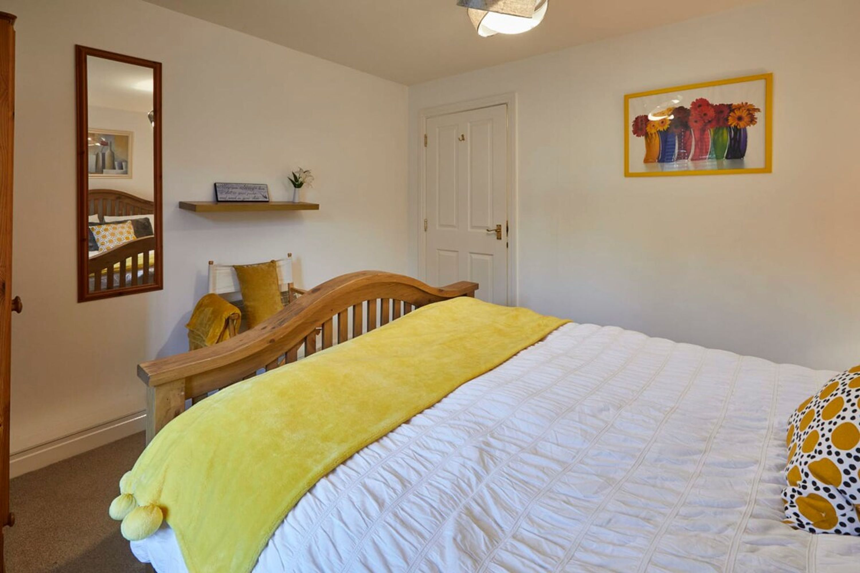 Une chambre à coucher avec un lit, une tête de lit en bois, un plaid jaune, une couette blanche, un miroir, une étagère avec une plante et un tableau représentant des fleurs accroché au mur.
