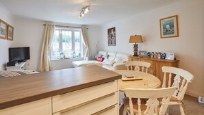 Un salon chaleureux avec un canapé, une table à manger et des chaises. On y trouve un téléviseur posé sur un meuble, une fenêtre avec des rideaux et une lampe sur une table d’appoint.