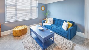 A living room with a blue sofa, a round ottoman, a coffee table, and a large window with blinds. - Host Stay Marshlands (Redcar)