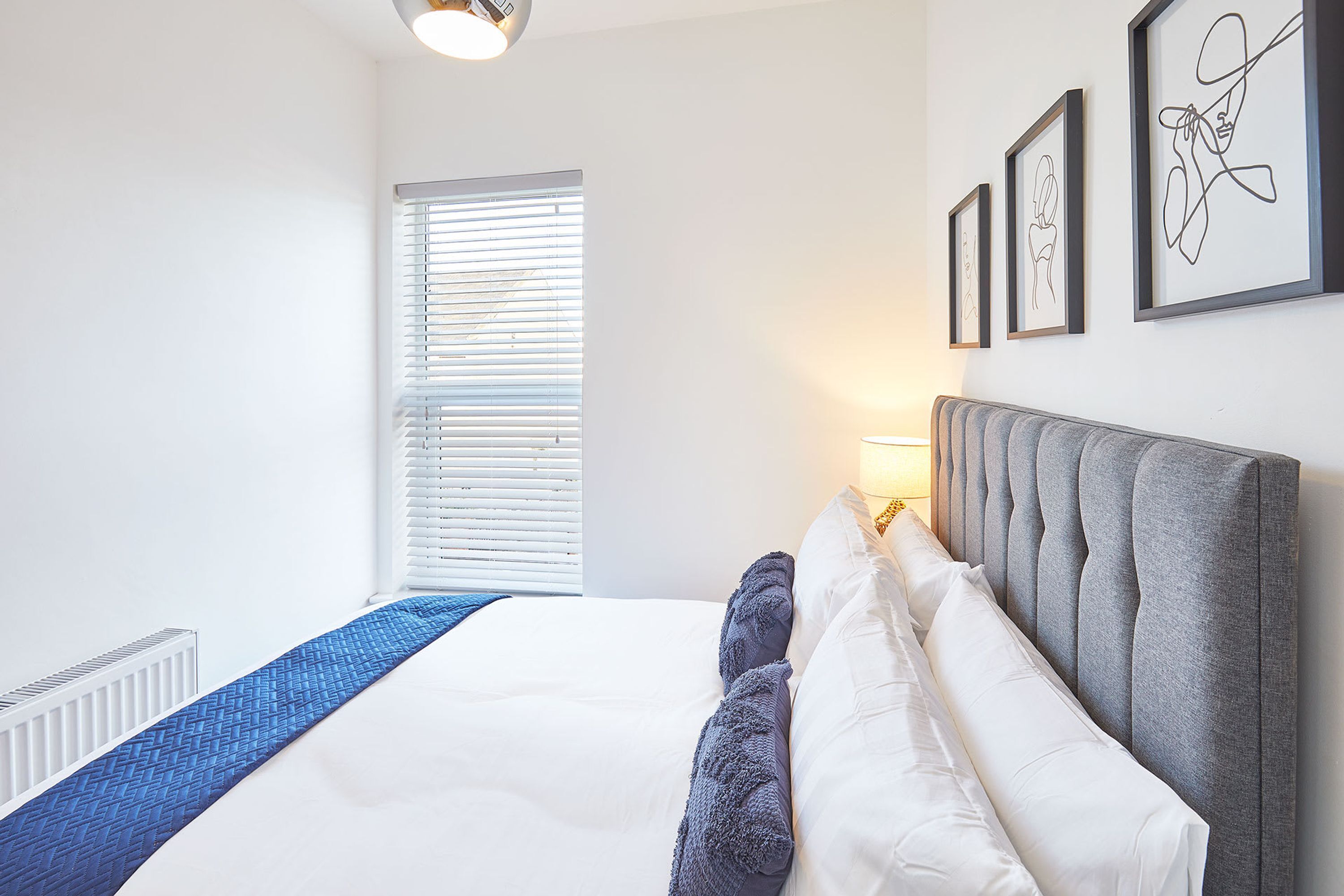 A neatly arranged bedroom with a large bed, a window with blinds, and three framed artworks on the wall.