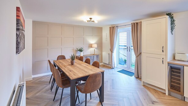 A dining area with a wooden table and chairs, a radiator, a window with curtains, and a kitchen area with cabinets. - Host Stay Redsky House (Whitby)