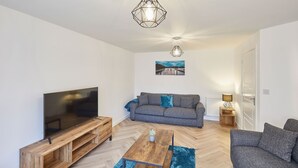 A modern living room with a flat-screen TV, wooden entertainment unit, grey sofa, blue rug, and two pendant lights. - Host Stay Redsky House (Whitby)