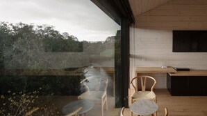 A modern interior with a large window overlooking a natural landscape. Inside, there are wooden chairs and a marble table.