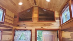 A wooden cabin with a loft bedroom, two windows with curtains, and a door. - Pleasant Grove Tiny Home! (Pell City)