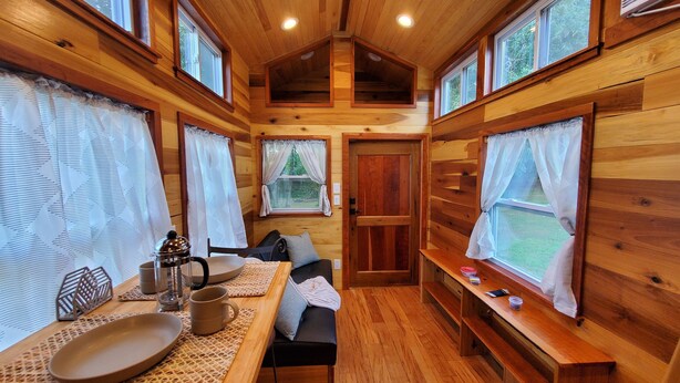 A cozy cabin interior with wooden walls, a dining table with chairs, a sofa, and a bench. - Pleasant Grove Tiny Home! (Pell City)