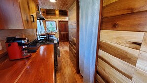 A wooden cabin interior with a kitchen area featuring a red stove, a dining table with chairs, and a view of the outdoors through a window. - Pleasant Grove Tiny Home! (Pell City)