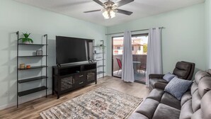 A living room with a flat-screen TV, dark wooden entertainment unit, a ceiling fan, a brown leather sofa, a patterned rug, and a sliding glass door leading to an outdoor area. - 1 Bedroom Condo, Sparkling Pool, Fitness Center, Pets Welcome (Glendale)